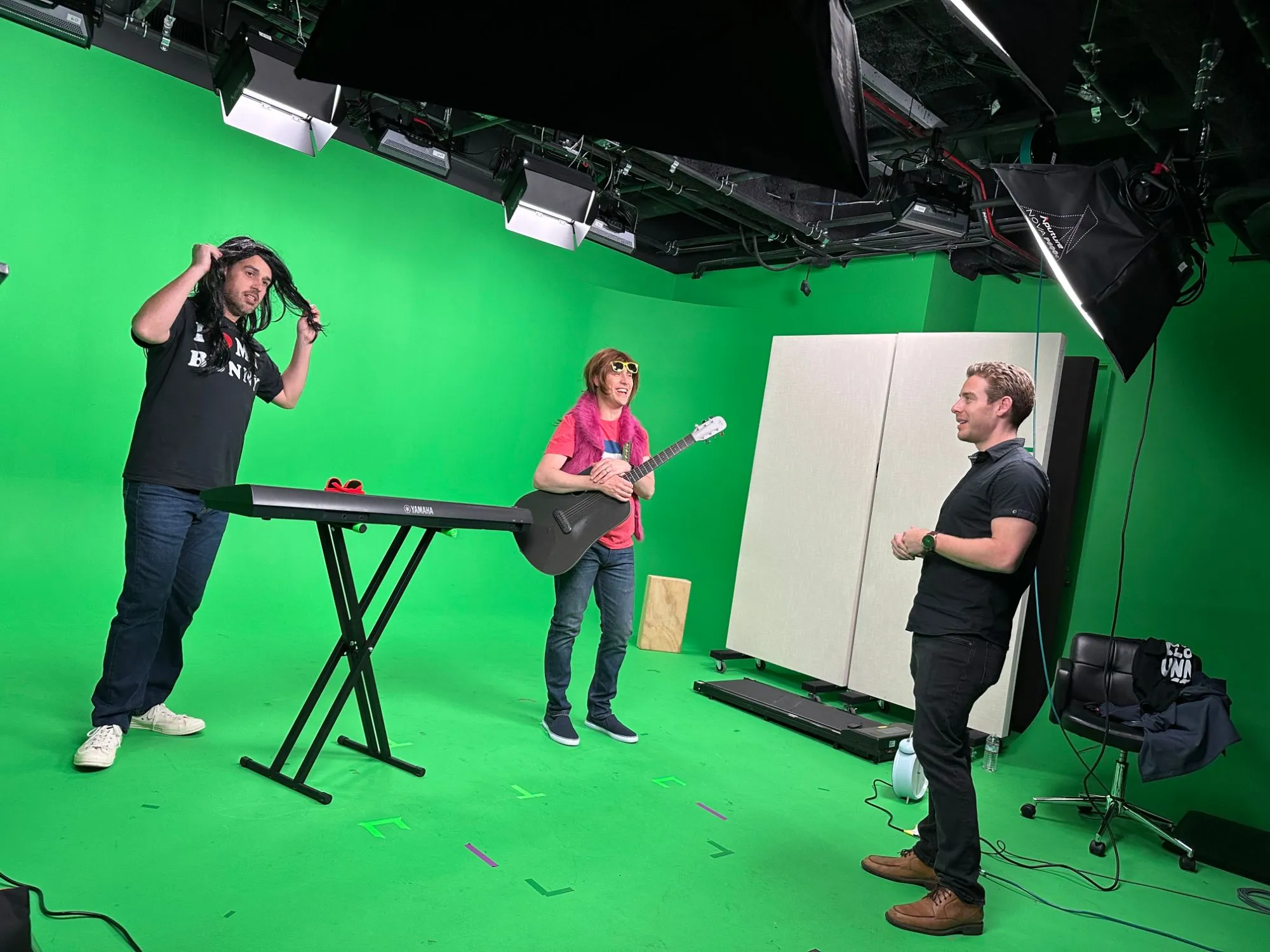 Two musicians in wigs with a keyboard and a guitar on a green-screen stage, with a crew member looking on