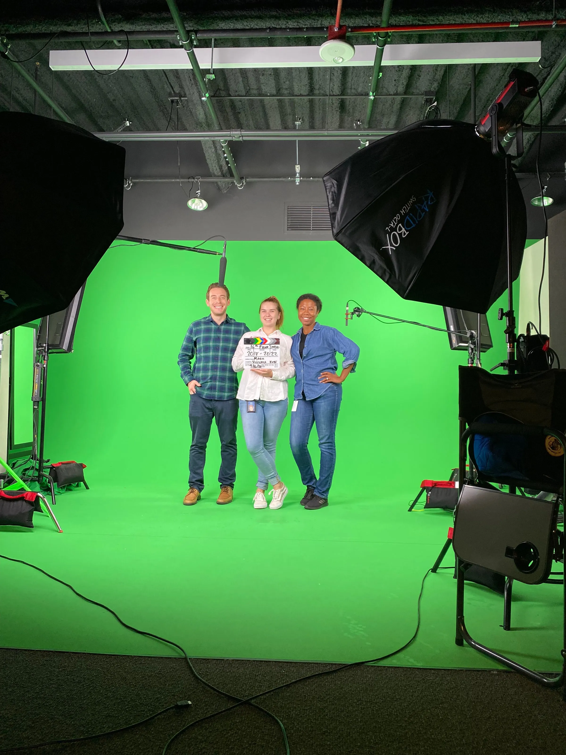 Three presenters standing on a lit green-screen stage between softboxes
