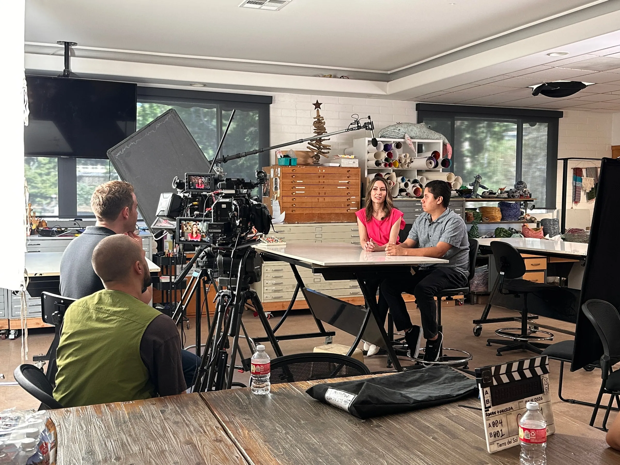 A two-person interview being filmed at a table in a classroom, with the camera operator and a boom in the foreground